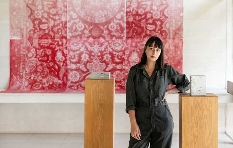 The artists Stanislava Pinchuk stands with two wooden plynths on either side of her. On the plynths are cubic sculptures. In the background, a red rug hangs from the wall. 