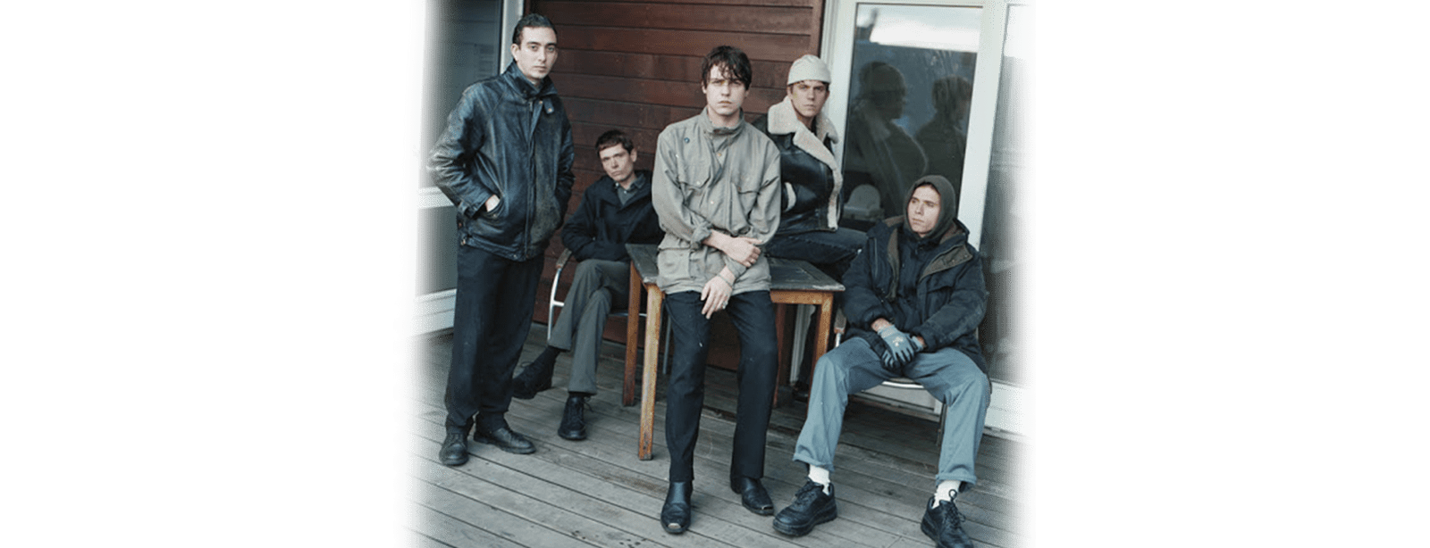 The band Iceage have their photo taken on the porch of a house. They stand or sit around a table dressed in muted colours, most staring into the camera.