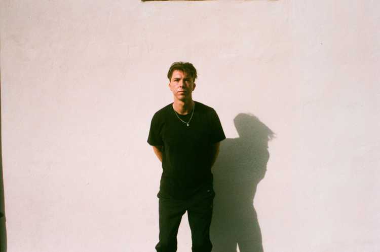 Self portrait of Calum Glendinning-Clark standing up against a sparse white wall on a sunny day. His expression is neutral and his hands are behind his back. Calum's shadow is dark and prominent on the wall.