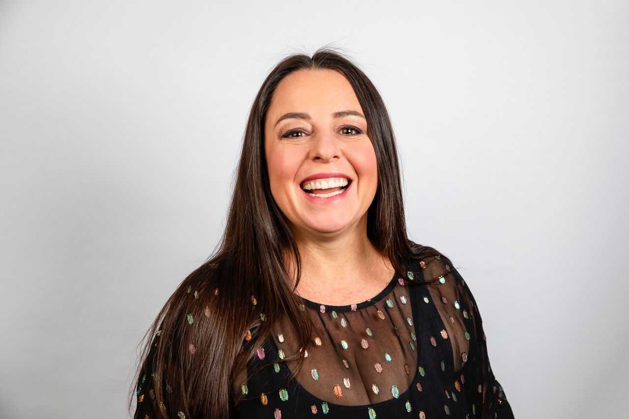 A photo of Myf Warhurst smiling in a brown dress with a blue background 