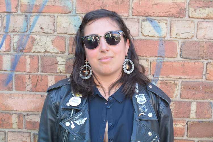 Photo of Christina wearing a black leather jacket, dark blue shirt and sunglasses, standing in front of a brick wall