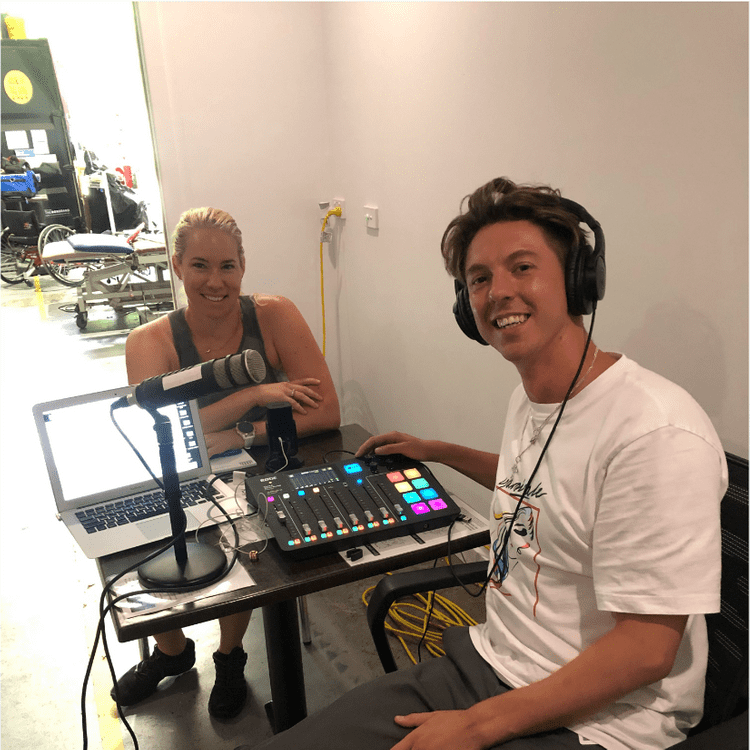 This is a photo of Calum and Pip who are posing for a photo just after their interview at Breaking Boundaries. Pip is blonde, smiling and wearing gym clothes. She looks directly at the camera. Calum is sitting opposite her, wearing a white t-shirt and grey slacks. He has headphones on and his hand is poised over the Rodecaster Pro
