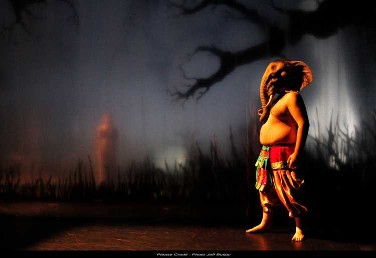 Two figures stand in a night scene on a theatre stage. The smaller figure is in the distance, behind long grass and a tree. The larger figure is a representation of Ganesh with a human body and an elephant’s head. 